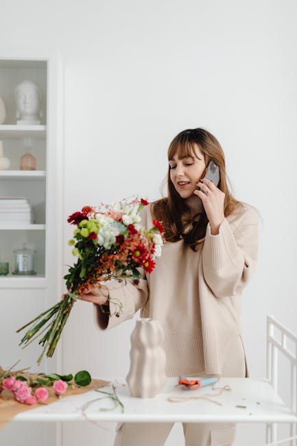 A woman with long brown hair and bangs, wearing a cream-colored sweater, is holding a vibrant floral bouquet in her left hand while speaking on a mobile phone with her right hand. The bouquet includes