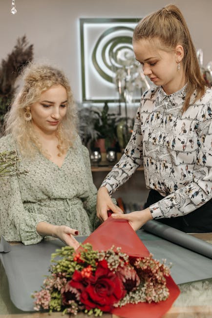 Two women are preparing a floral gift at a table in a well-lit interior space. The older woman has wavy, blonde hair and wears a light green, patterned dress, while the younger woman has straight, lig