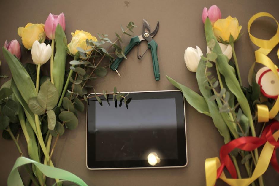 A flat-lay image of a floral arrangement workspace featuring partially assembled bouquets of pink, yellow, and white tulips with green stems and leaves, arranged on a neutral-colored surface. In the c