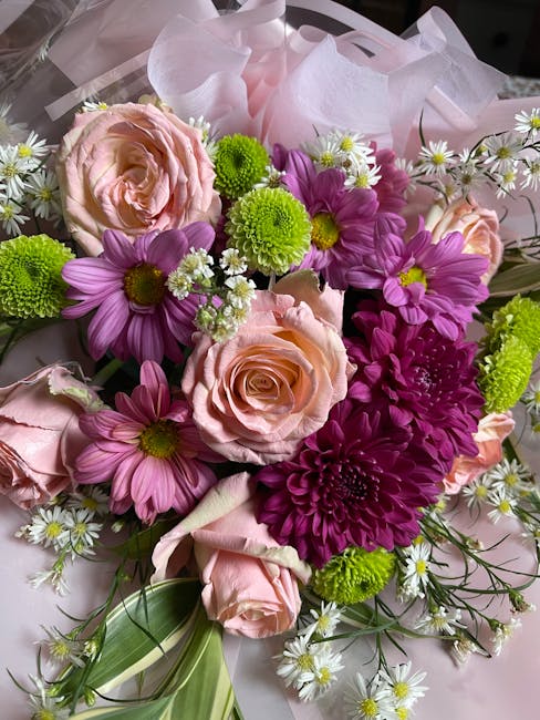 A vibrant floral arrangement featuring soft pink roses with layered petals and a gentle blush hue, complemented by bright purple daisies with yellow centers, fresh green chrysanthemum-like blooms, and