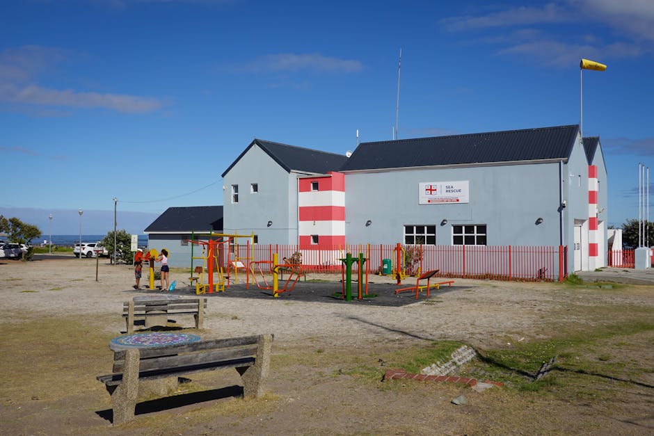 A light blue, two-story Sea Rescue building with red and white striped accents on the corners and a white sign with red and blue markings, located in a coastal area under a clear blue sky. In front of