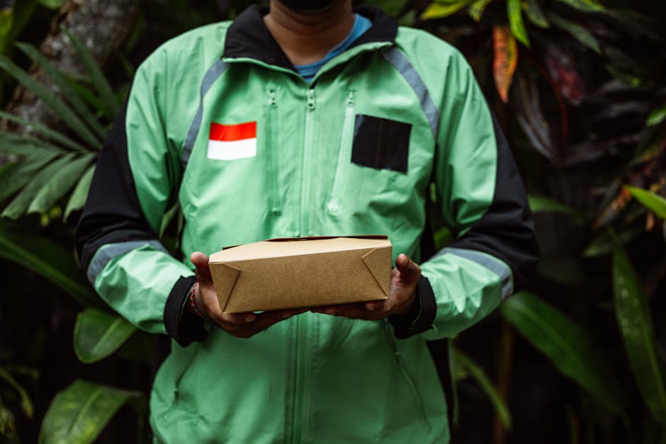 A delivery person wearing a green and black jacket with a red and white flag patch on the chest is holding a rectangular cardboard box, presumably containing flowers, in front of lush green foliage in