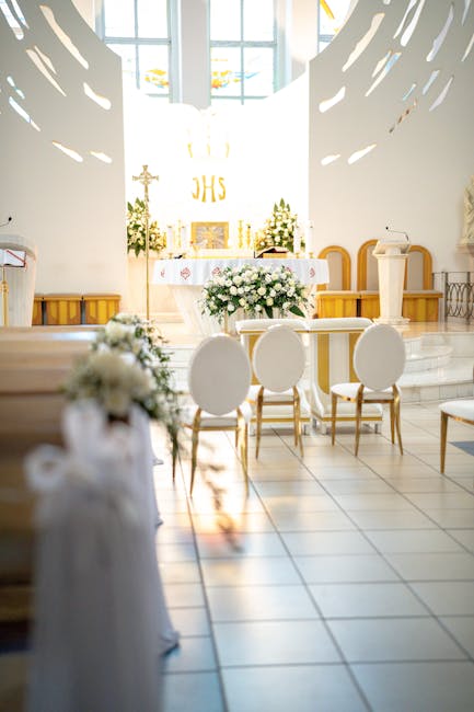 A wedding ceremony setup inside a modern, bright church with white walls and contemporary design elements. In the foreground, a floral arrangement featuring white roses, hydrangeas, and greenery is pl