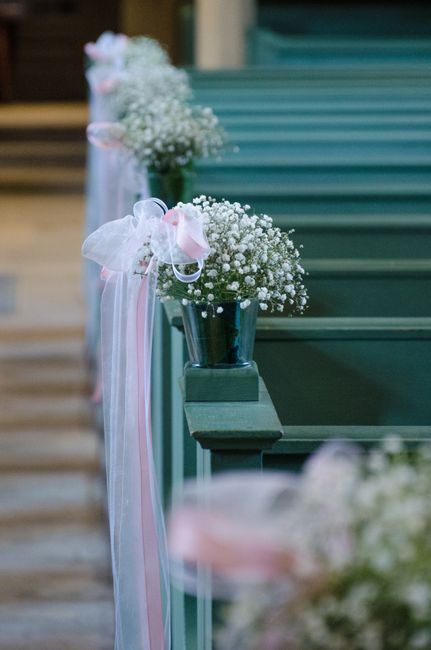A row of elegant flower arrangements consisting of small white baby's breath blooms in metallic vases, placed on dark green wooden pews or benches, with delicate pastel pink and white ribbons tied to 
