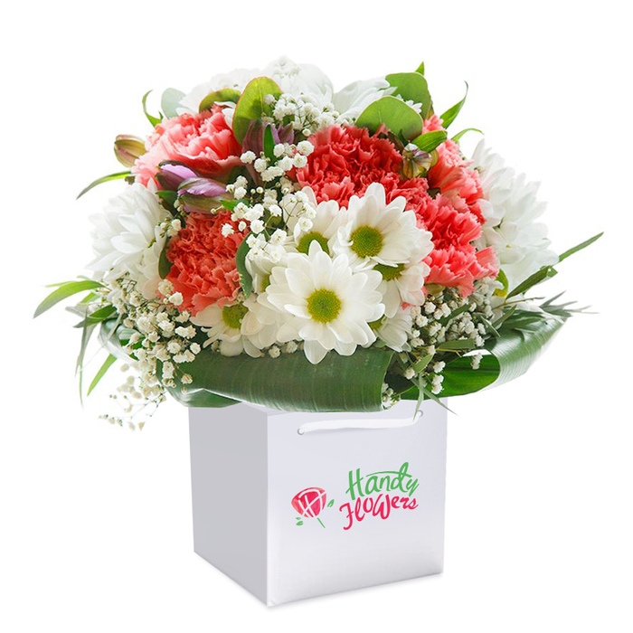 Resting upright in a flawless white square gift bag, this luxurious floral arrangement is a true showcase of expert floristry in Rickmansworth. The bouquet forms a seamless blend: ruffled carnations in soft coral, light peach, and lively red arrayed with natural elegance, each petal layered to create visual depth and tactile richness. Among these, clusters of bright white daisy-like chrysanthemums with vivid lime-green centres provide a crisp, contemporary edge-echoing the modern charm found along Rickmansworth High Street. Sprigs of snowy baby's breath are woven throughout, their airy clusters evoking gentle morning fog rising over Batchworth Lake. At the periphery, the composition is framed by bold, deep green foliage, their glossy finish cradling the blooms like a lush green collar. A few closed buds edged in purple and blush peek from within, offering hints of potential and renewal, much like the quiet promise of emerging spring in local parks. The gift bag, sturdy and designed for easy transport, features pristine white ribbon handles-ready to surprise a loved one near Rickmansworth Station or delivered to a local business as a thoughtful gesture. The white backdrop amplifies the bouquet's vibrant tones, making every flower stand out and capturing the authentic freshness that customers trust from their local florist. This arrangement radiates thoughtfulness-perfect for birthdays, anniversaries, or to add a touch of seasonal beauty to any Rickmansworth home.
