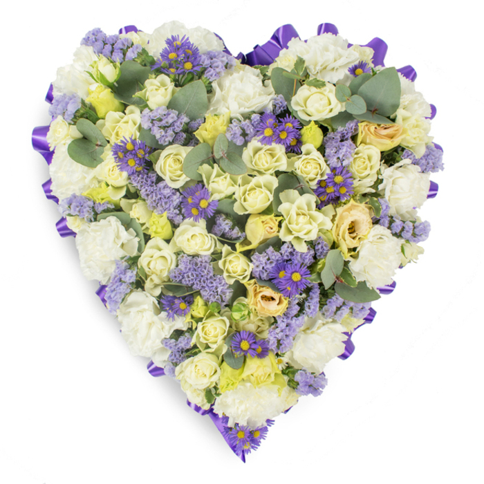 A deeply considered heart-shaped wreath photographed against a crisp white backdrop, this memorial arrangement blends traditional sympathy flowers with contemporary texture to create a serene, dignified tribute. The dense composition is dominated by creamy and pale yellow roses, their varied stages of bloom providing soft, velvety depth and a comforting lift; interspersed white carnations add generous body and a time-honoured tenderness familiar at wakes and services. Delicate sprays of lavender-coloured statice weave through the blooms like gentle echoes of remembrance, while small purple daisy-like asters with bright yellow centres give lively, soulful highlights that draw the eye inward. Cool, rounded eucalyptus leaves - blue-green in tone and matte in finish - are tucked throughout to punctuate the massed petals and to introduce a subtle, leafy aroma by association. Encircling the entire heart is a rich purple satin ribbon, looped and laid with exacting care to produce a polished, refined border that holds the composition together. The studio lighting accentuates petal texture and colour gradation without glare, presenting an image of calm and thoughtful presence. Prepared by local hands familiar with the quiet needs of Rickmansworth families, from High Street vigils to chapel services near Batchworth Lock, this arrangement offers a respectful way to convey sympathy and lasting affection. Flowers Rickmansworth crafts each piece with freshness and care, making this heart a fitting symbol of sorrow, devotion, and remembrance.