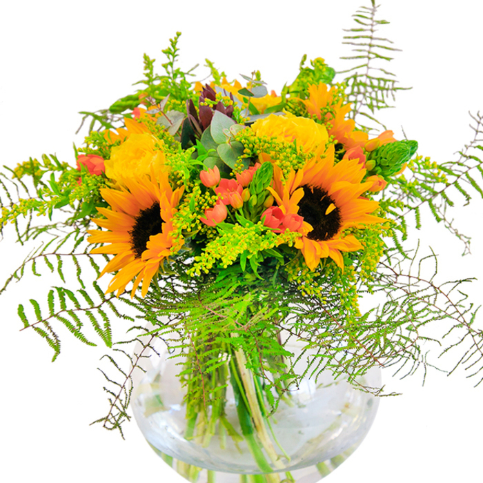 The moment your gaze lands on this brilliant bouquet, you're greeted by the vibrant energy of sunflowers and roses in full bloom-a scene reminiscent of Rickmansworth's finest gardens flourishing by Maple Cross. Sunflowers, large and radiant, anchor the design with dark, earthy centers and petals that blaze with hues of yellow and orange, breathing sunshine and happiness into the arrangement. Just beside, yellow garden roses unfold in intricate, spiraling layers, their fragrant, creamy petals providing a rich textural contrast and a touch of timeless grace, much like a classic Rickmansworth afternoon tea. Interspersed throughout are dazzling Hypericum berries, each cluster gleaming crimson against a backdrop of deep and seasonal greens. Fern fronds and light, feathery filler spill gently from the bouquet, echoing the wild, untamed charm of local woods near Stocker's Lake, while the slender, upright greens and bluish eucalyptus add a refreshing, botanical accent. The clear, round vase cradles each stem, its transparency offering glimpses of water shimmering in the morning sun, tying the bouquet together with a sense of clarity and natural elegance. The bouquet releases a subtle, uplifting fragrance-delicate yet persistent, reminiscent of open-air walks near Rickmansworth's leafy parks. Perfect for commemorating joyous occasions, this arrangement brings the energy of a summer celebration and a hint of local artistry into any space it graces.